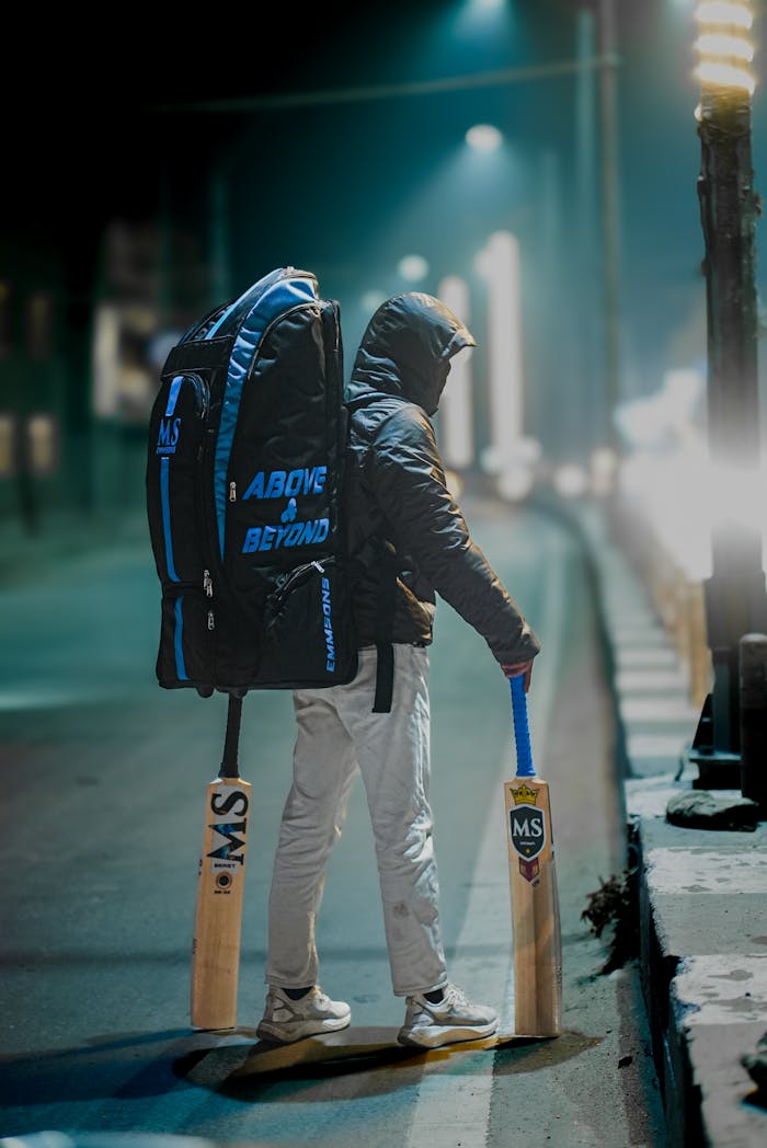 A young cricketer with gear stands on a dimly lit street at night holding bats.